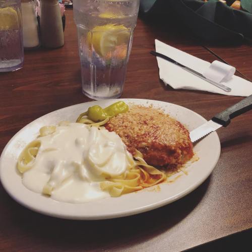 A plate of chicken parmesan with fettuccine alfredo and green peppers, alongside a glass of water with lemon.