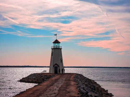 A lighthouse stands on a rocky pier, with a colorful sky and calm water in the background.