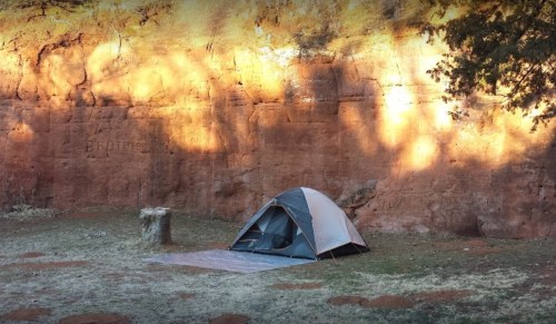 A tent set up on grass near a red rock wall, with soft lighting creating shadows in the background.