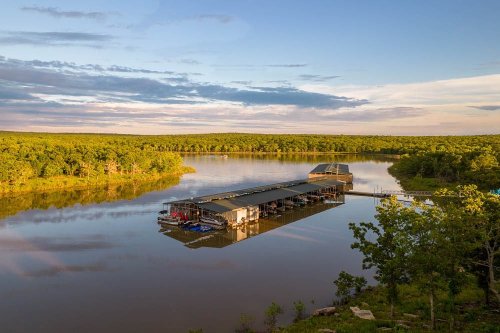 A serene lake scene with a dock and boats, surrounded by lush green trees under a partly cloudy sky.