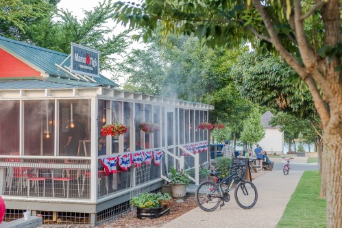 A cozy restaurant with outdoor seating, decorated with flowers and patriotic banners, surrounded by trees and a bike.