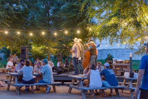 A lively outdoor scene with musicians performing on stage, surrounded by people enjoying food and drinks at picnic tables.