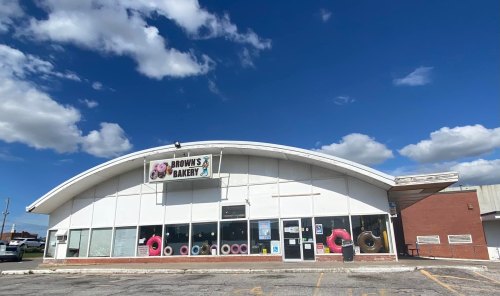 A bakery with a curved roof, large windows, and colorful donut decorations on display, set against a blue sky.