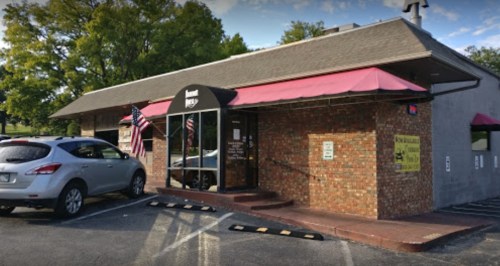 A brick building with a red awning, featuring a sign and American flags outside, parked cars nearby.