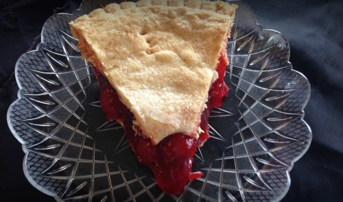 A slice of cherry pie on a decorative glass plate, with a golden crust and vibrant red filling.