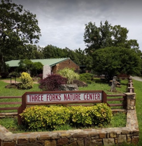 A sign for Three Forks Nature Center in a landscaped area with trees and a building in the background.