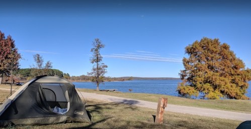 A tent by a serene lake, surrounded by trees and a clear blue sky, with a path along the water's edge.