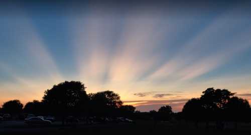 A serene sunset with colorful rays spreading across the sky, silhouetting trees and a distant parking area.