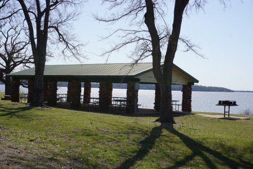 A lakeside pavilion with stone columns, surrounded by trees and picnic tables, overlooking the water.