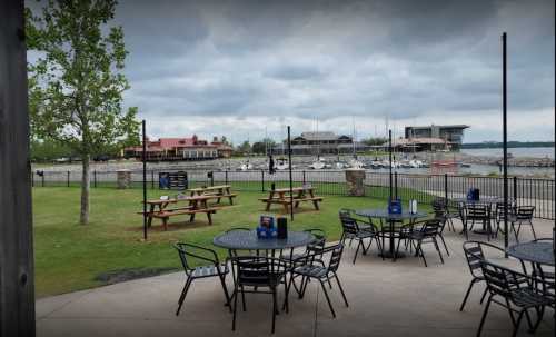 Outdoor dining area with tables overlooking a marina and cloudy sky, featuring boats and a restaurant in the background.