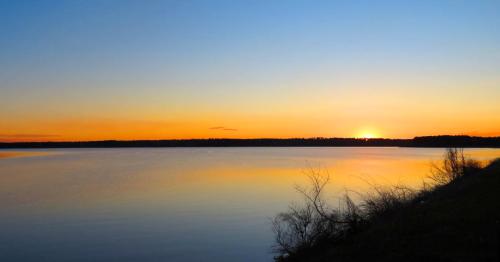 A serene lake at sunset, with vibrant orange and blue hues reflecting on the calm water and silhouetted trees along the shore.
