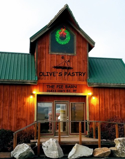 A rustic wooden building with a green roof, featuring a festive wreath and signs for "Olive's Pastry" and "The Pie Barn."