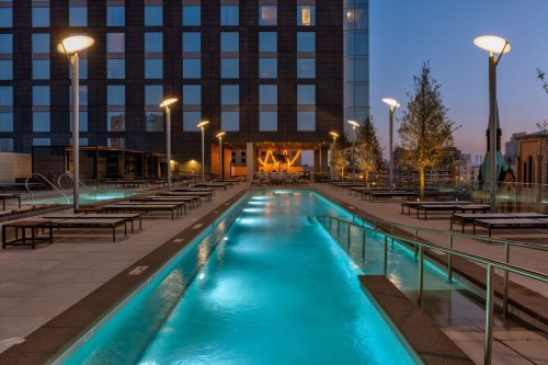 A modern rooftop pool area with illuminated water, lounge chairs, and a city skyline in the background at dusk.