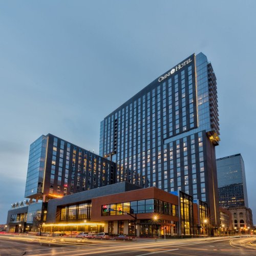 Modern hotel building with large glass windows, illuminated at dusk, surrounded by city streets and traffic.