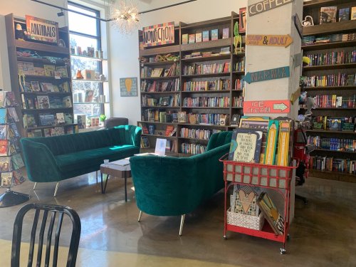 Cozy bookstore interior with green sofas, bookshelves filled with books, and a cart displaying colorful items.