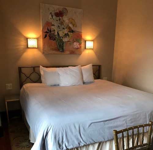 A cozy bedroom featuring a bed with white linens, a floral painting, and soft wall lighting.