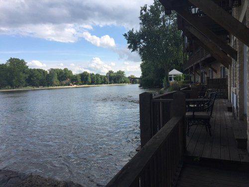 A serene river view with trees lining the banks and a wooden deck with chairs in the foreground under a cloudy sky.