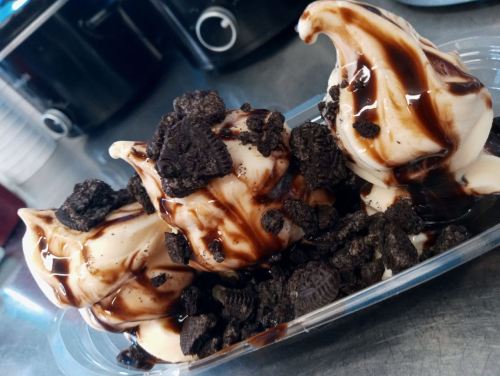 A dessert cup with three swirls of soft serve ice cream topped with crushed Oreos and drizzled with chocolate sauce.