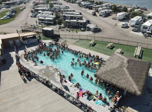 Aerial view of a crowded pool area with people swimming, surrounded by RVs and a thatched-roof cabana.