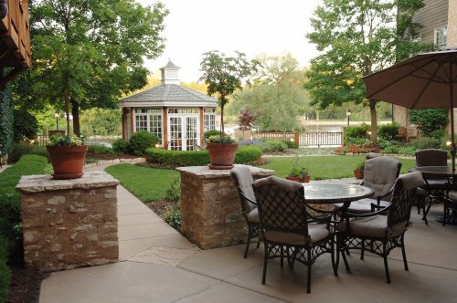 A serene garden with a gazebo, lush greenery, and a patio set, overlooking a calm body of water.