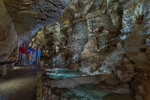 A group of visitors explores a cavern with rock formations and clear water pools, illuminated by soft lighting.