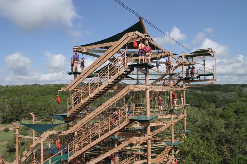 A multi-level outdoor ropes course with participants on platforms and zip lines, surrounded by trees and blue skies.