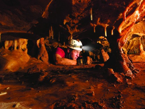 A caver in a helmet explores a narrow cave passage, illuminated by a headlamp, surrounded by stalactites and rock formations.