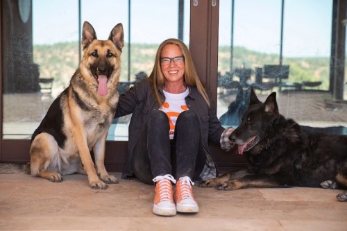 A woman with glasses sits on the floor between two dogs, a German Shepherd and a black dog, in a bright indoor space.