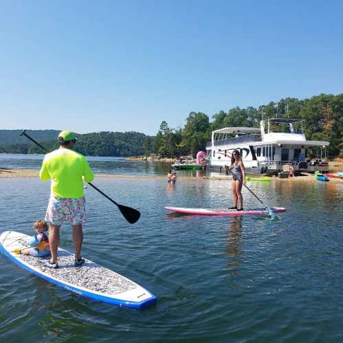 A man paddleboards with a child, while two others paddleboard nearby, with a boat and trees in the background.