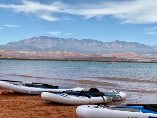 Kayaks lined up on a sandy beach with mountains and a calm lake in the background under a clear blue sky.