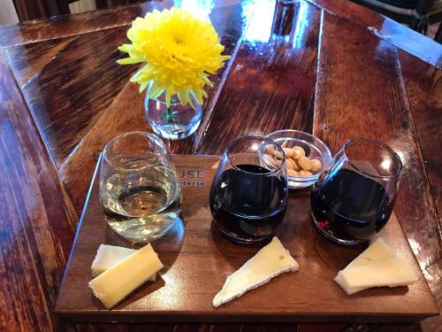 A wooden platter with two glasses of wine, cheese slices, nuts, and a yellow flower in a vase on a wooden table.