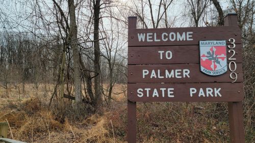 Wooden sign reading "Welcome to Palmer State Park" with trees and bare ground in the background.