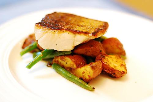 A plate of seared fish atop roasted potatoes and green beans, garnished and presented elegantly.