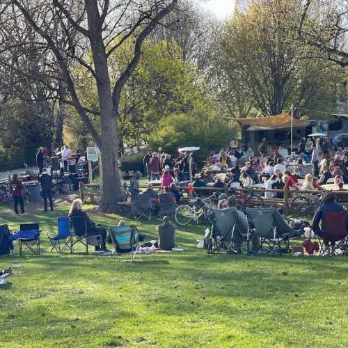 A lively outdoor gathering in a park with people sitting on blankets and chairs, enjoying food and music under trees.