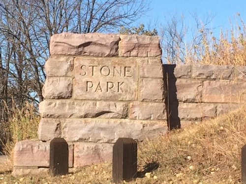 A stone sign reading "STONE PARK" stands on a grassy area with trees in the background under a clear blue sky.