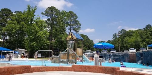 A sunny pool area featuring a water slide, umbrellas, and people enjoying the water surrounded by trees.