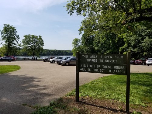 A park area with a sign about open hours, surrounded by trees and a parking lot near a lake.