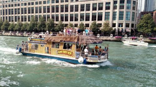 A party boat with a thatched roof cruises on a river, filled with people enjoying the sunny day.