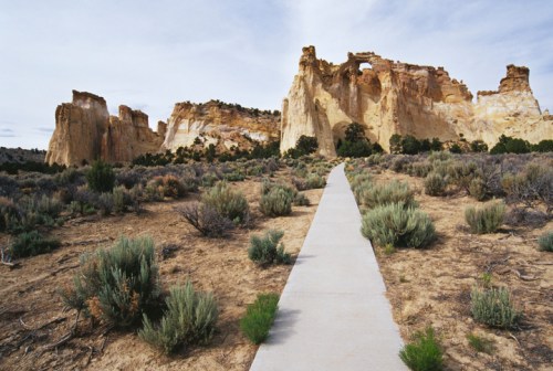 A paved path leads through a desert landscape with towering rock formations and sparse vegetation under a cloudy sky.