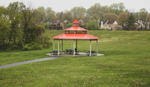 A red-roofed gazebo with picnic tables sits in a grassy area, surrounded by trees and residential buildings in the background.
