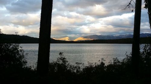 A serene lake surrounded by trees, with mountains in the background under a cloudy sky. Soft light illuminates the peaks.