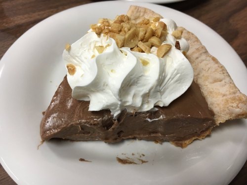 A slice of chocolate cream pie topped with whipped cream and chopped nuts on a white plate.