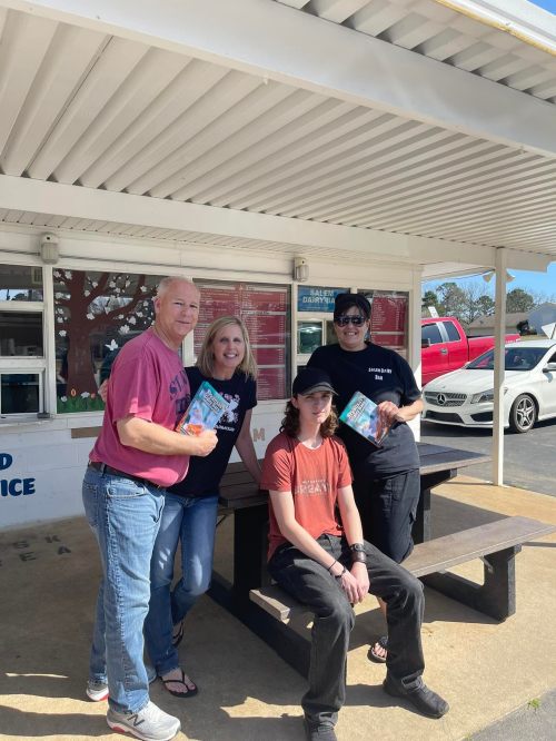 Four people pose together outside a building, holding books and smiling, with a picnic table in the foreground.