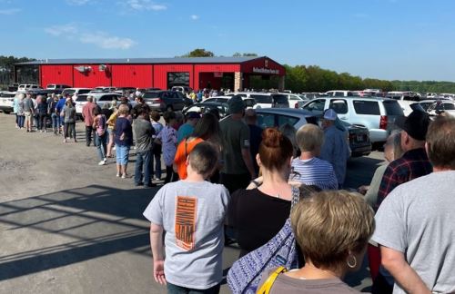 A long line of people waiting outside a red building, with cars parked nearby under a clear blue sky.