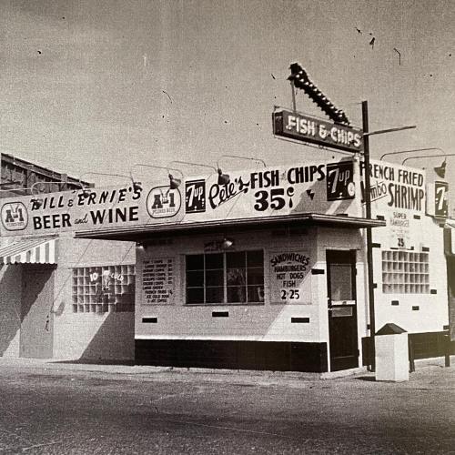 Black and white photo of a vintage fish and chips shop with signs for beer, wine, and sandwiches.