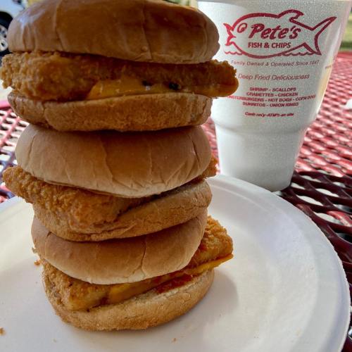 A stack of three fried fish sandwiches on a plate, next to a cup from Pete's Fish & Chips.