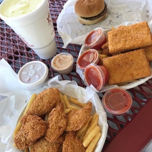 A tray of fast food featuring chicken nuggets, fries, a burger, and dipping sauces, alongside a large drink.