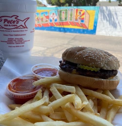 A plate of fish and chips with a burger, fries, and dipping sauces, set against a colorful mural background.