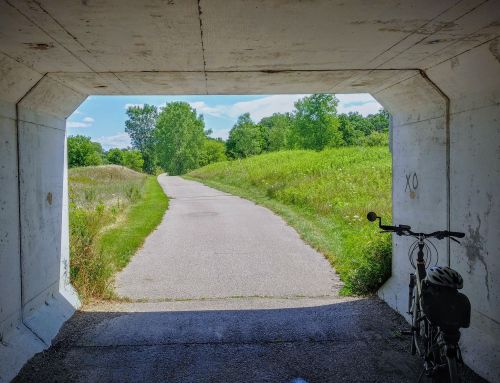A bike leans against a concrete tunnel, leading to a sunny path through green fields and trees.