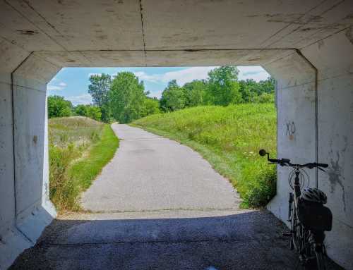 A bike leans against a concrete tunnel, leading to a sunny path through green fields and trees.